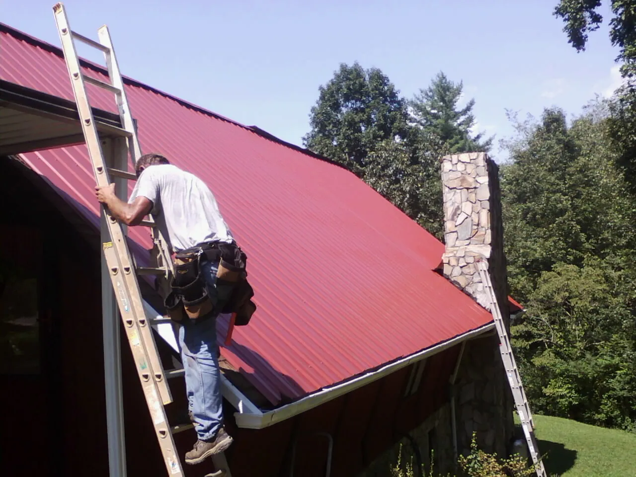 Roofer on a ladder beside a red metal roof and stone chimney — Soffit & Fascia Repair by Roofing Brothers in Malden